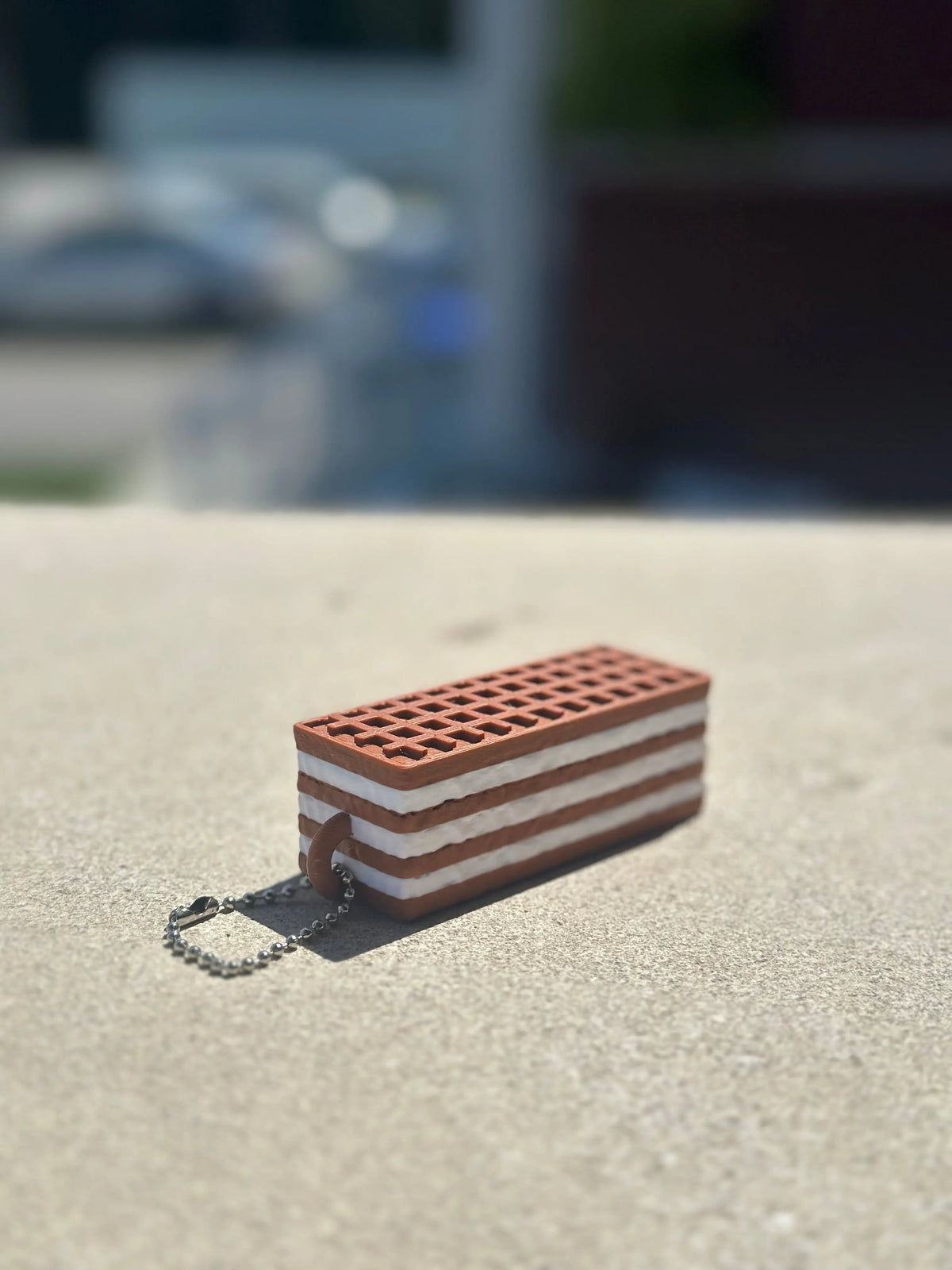 A small rectangular chocolate wafer biscuit-shaped fidget clicker keychain with a textured grid pattern and attached silver ball chain, photographed outdoors on a concrete surface with a blurred background.