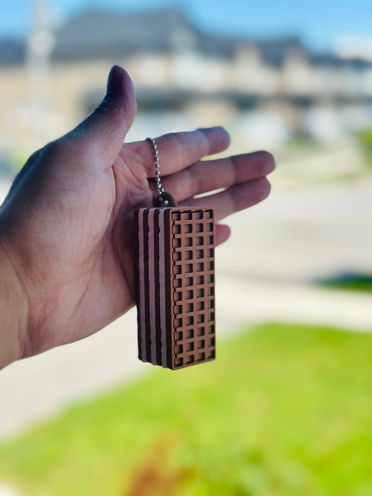 A hand holding a chocolate wafer biscuit-shaped fidget clicker keychain with a realistic waffle-textured surface and silver ball chain, displayed against a softly blurred outdoor background. The portable 3D-printed clicker is designed to resemble a layered chocolate wafer biscuit.