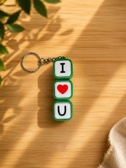 A keychain made of three stacked square tiles reading 'I' and 'U' with a red heart in the middle, attached to a metal key ring. The keychain is placed on a wooden surface with leaf shadows and warm sunlight.