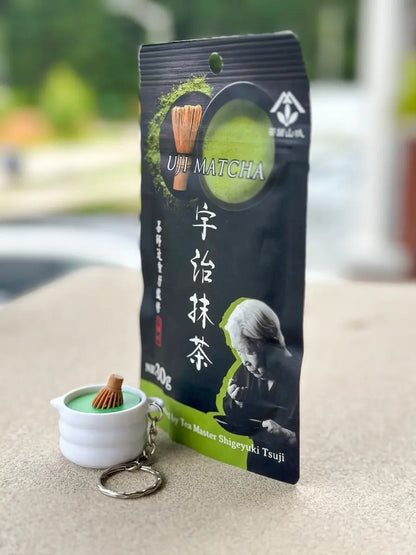 A 3D-printed matcha whisk clicker keychain sits beside a package of matcha tea on a light stone surface. The miniature whisk-shaped fidget features a tactile clicking mechanism and is displayed as a decorative accessory, with a metal keychain attachment visible nearby. The matcha tea package serves as a prop to showcase the product's matcha-inspired design.