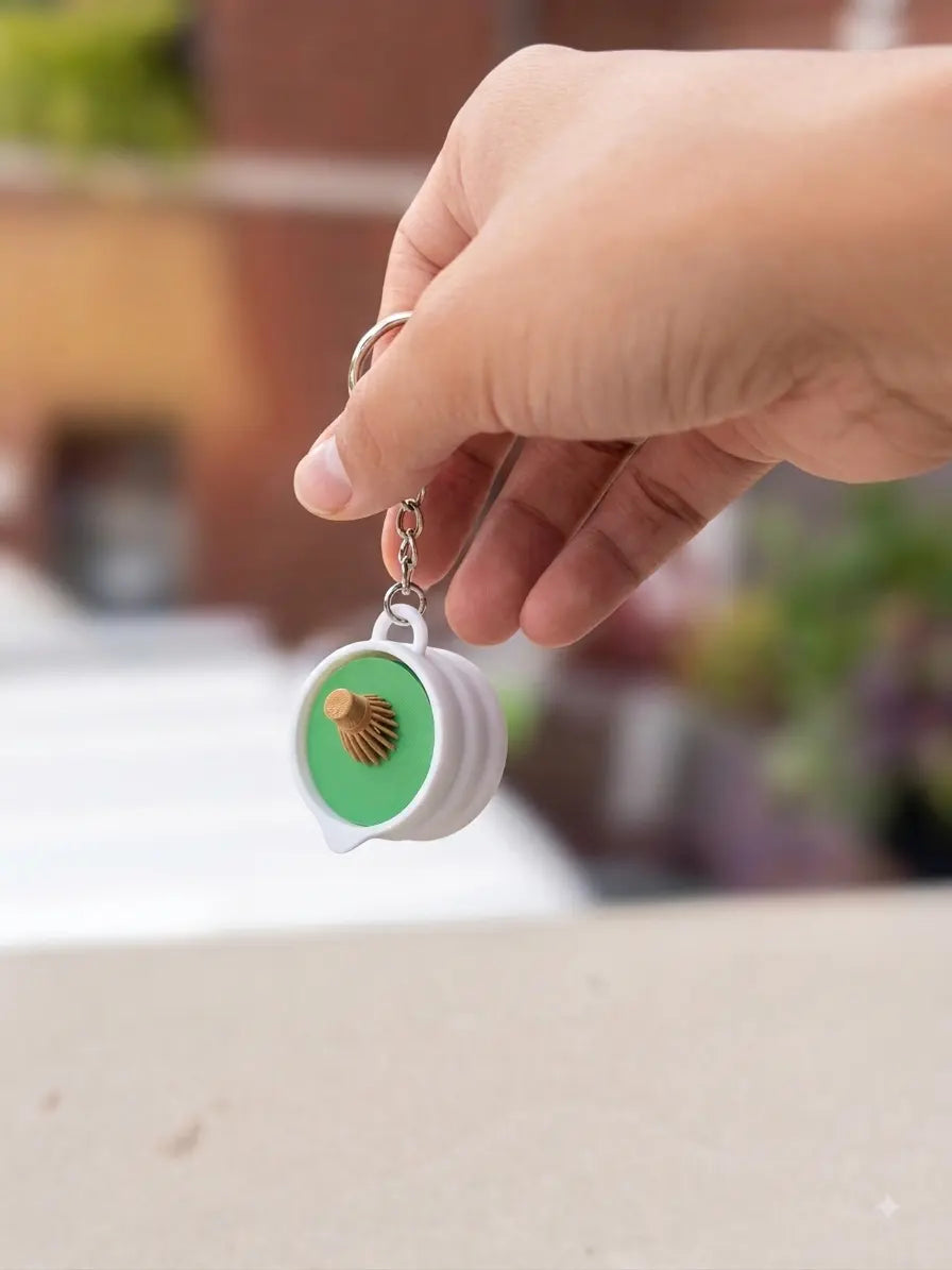A hand holds a miniature 3D-printed matcha whisk clicker keychain, inspired by traditional Japanese tea-making tools. The green-and-white accessory features a compact whisk-shaped design with a tactile clicking mechanism, displayed against a softly blurred outdoor background.