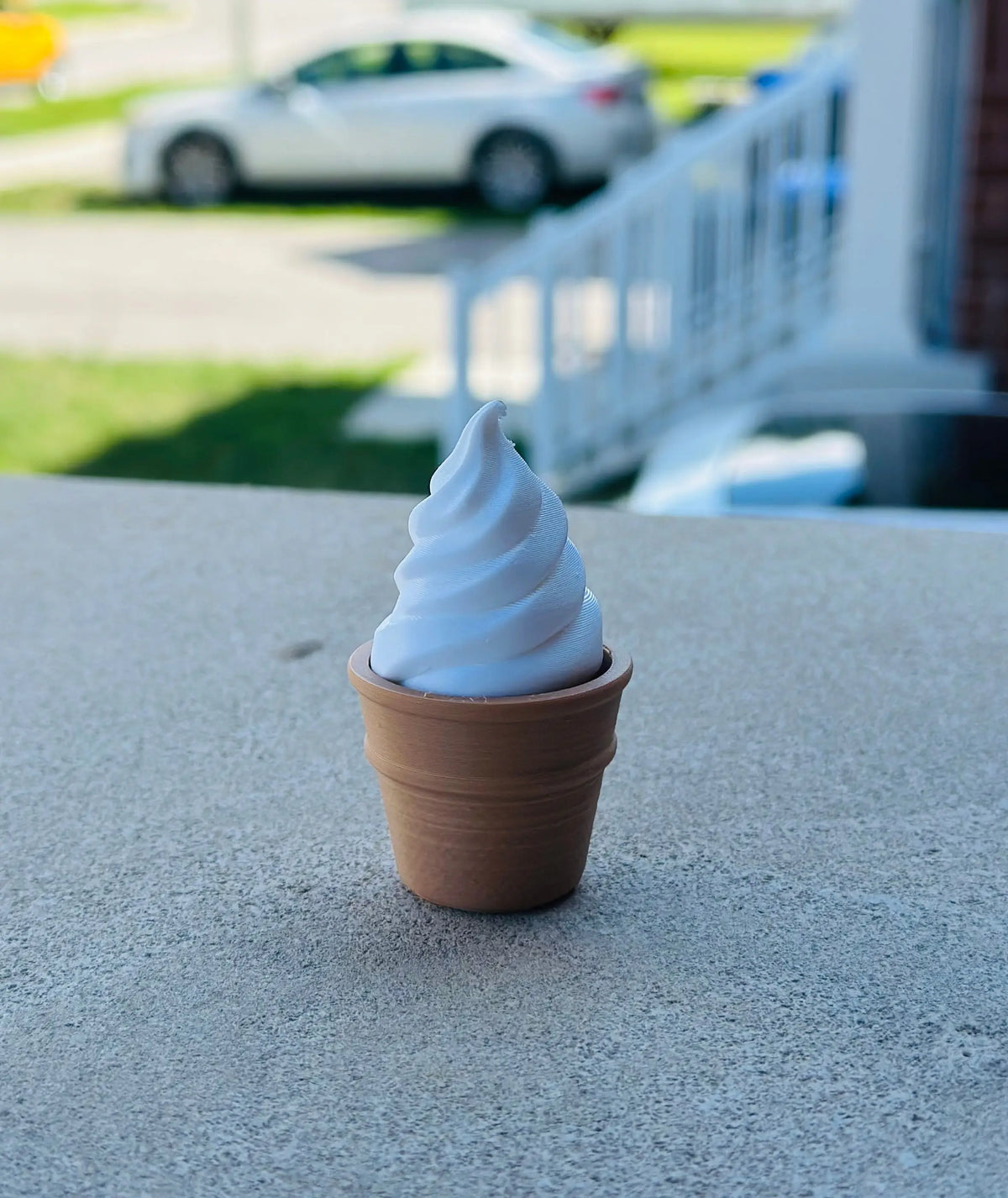 A 3D-printed vanilla soft-serve ice cream fidget clicker featuring a white swirl top in a brown cone, set on an outdoor concrete surface with a house and yard in the background.