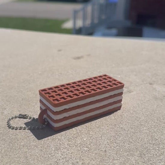 A chocolate wafer biscuit-shaped fidget clicker rests on a concrete surface while a finger repeatedly presses the clicker, demonstrating its satisfying clicking action.