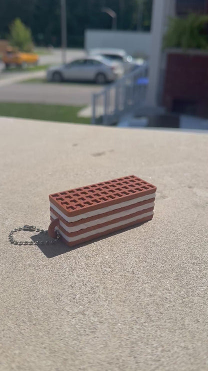 A chocolate wafer biscuit-shaped fidget clicker rests on a concrete surface while a finger repeatedly presses the clicker, demonstrating its satisfying clicking action.