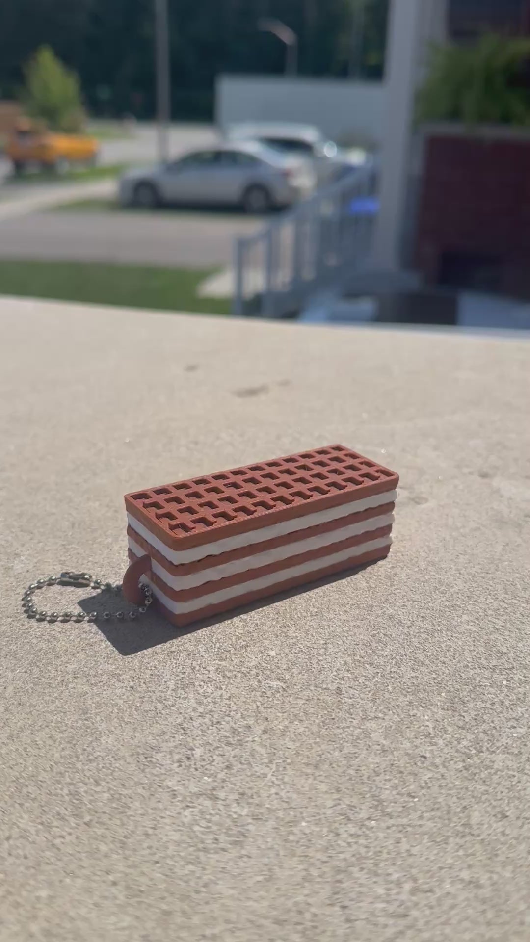A chocolate wafer biscuit-shaped fidget clicker rests on a concrete surface while a finger repeatedly presses the clicker, demonstrating its satisfying clicking action.